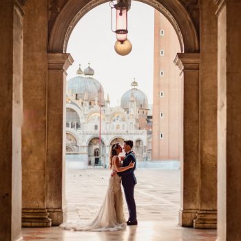 Couple under St. Mark’s square arcades with the basilica in the background – Venice Wedding Photography Engagement.