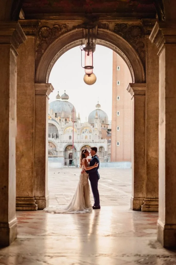 Couple under St. Mark’s square arcades with the basilica in the background – Venice Wedding Photography Engagement.