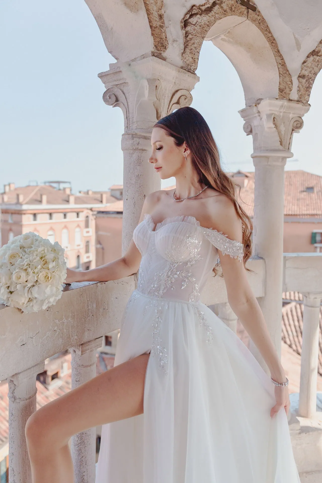 Venice wedding bride in elegant off-shoulder gown on historic balcony with city view.