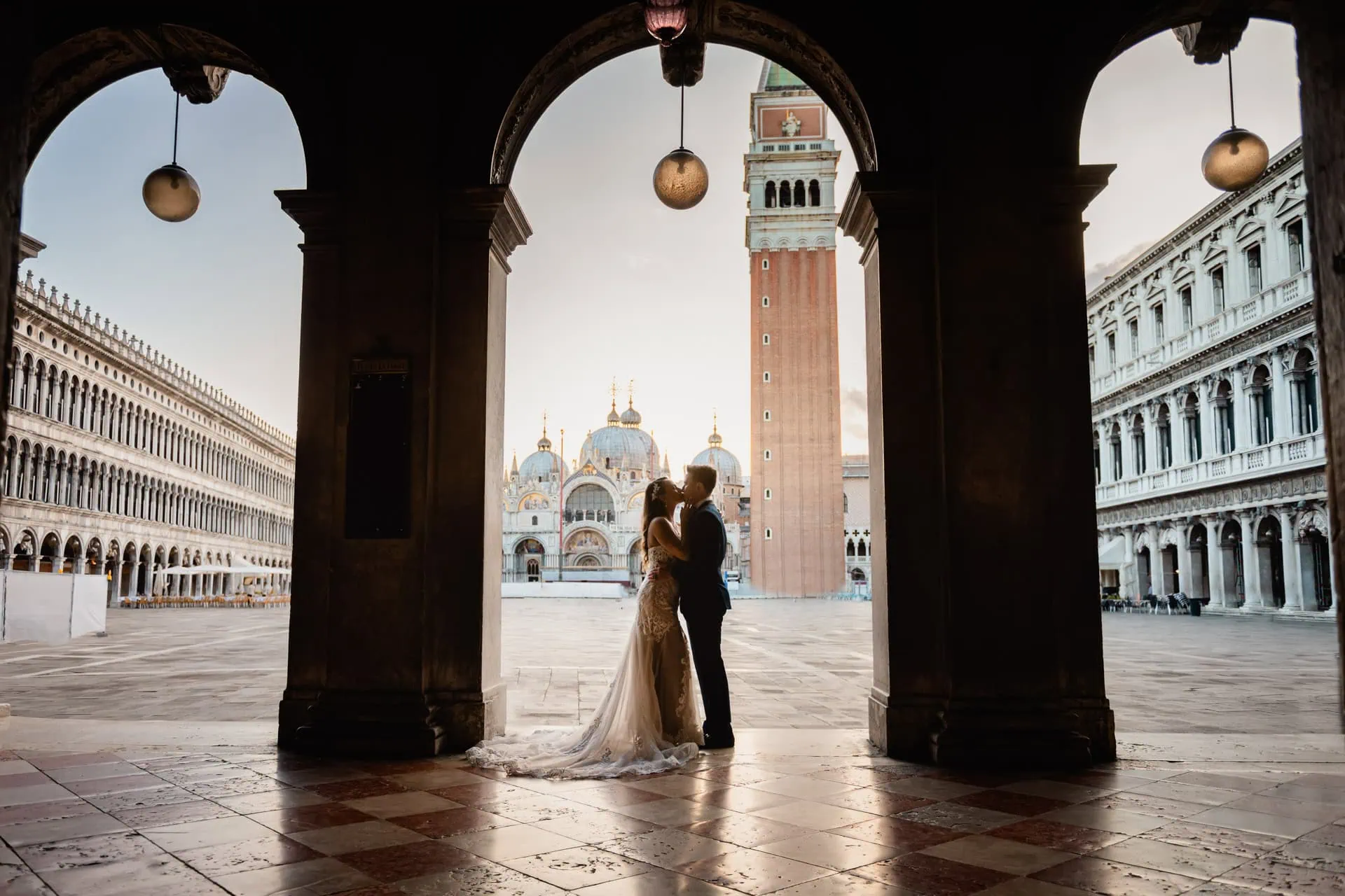 Bride and groom walk hand in hand through an empty Piazza San Marco, with the Bridge of Sighs in the background.