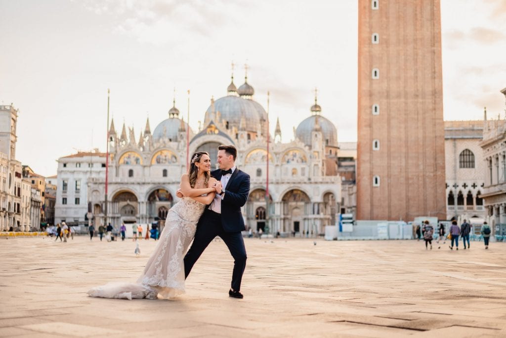 Bride and groom walk hand in hand through an empty Piazza San Marco, with the Bridge of Sighs in the background.