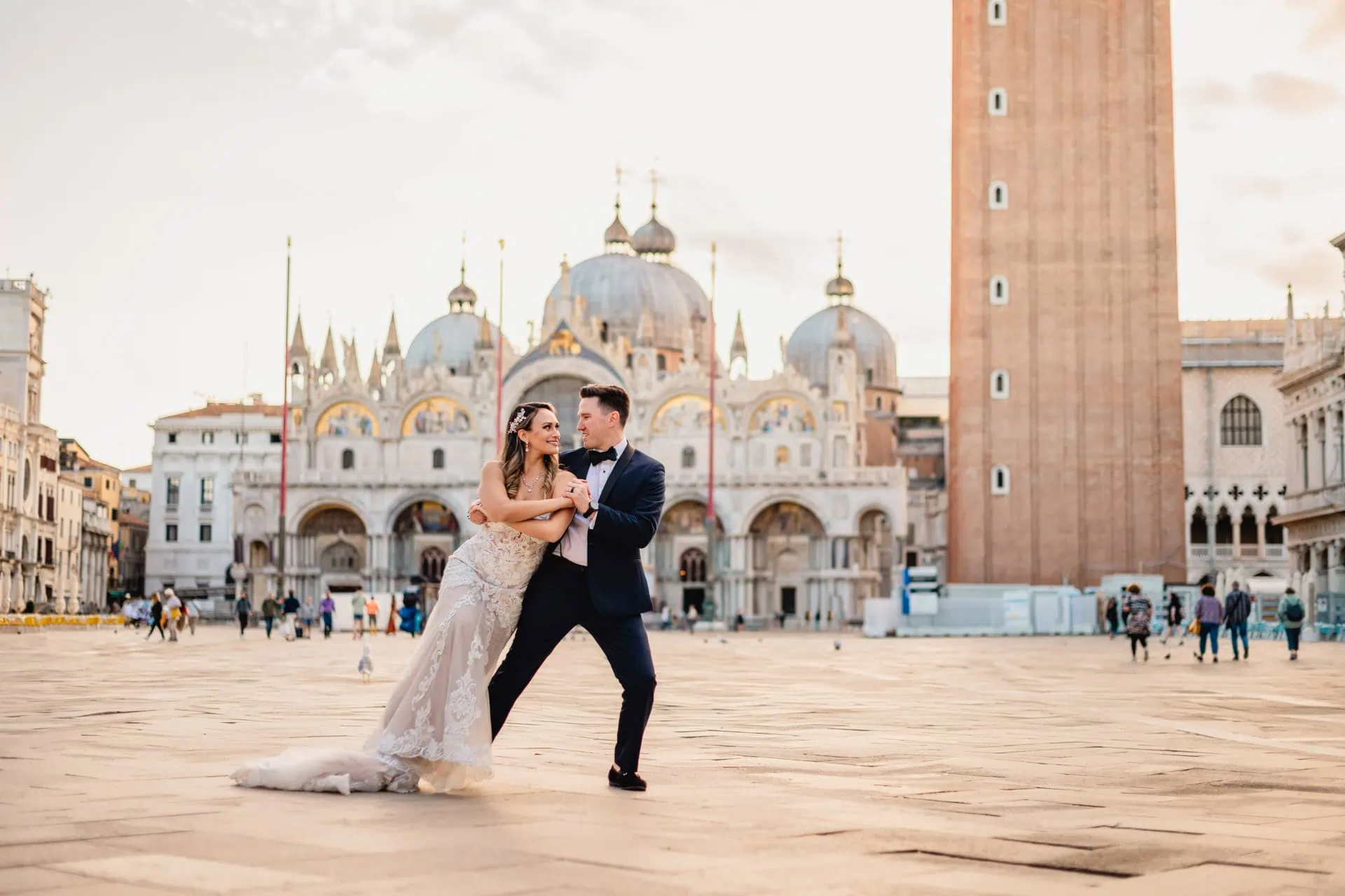 Bride and groom walk hand in hand through an empty Piazza San Marco, with the Bridge of Sighs in the background.