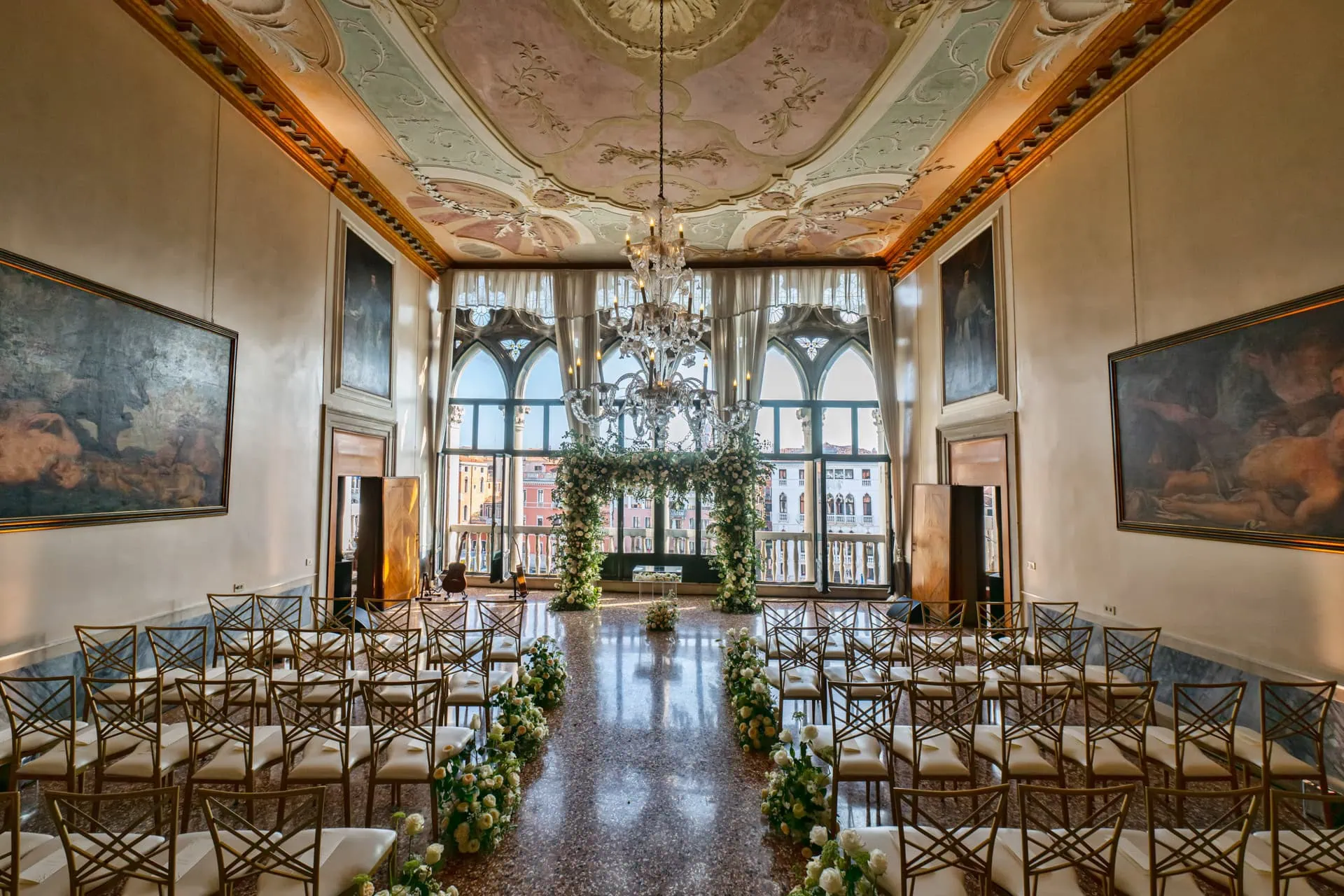 Elegant Venetian wedding ceremony setup with floral arch and chandelier in a historic hall.