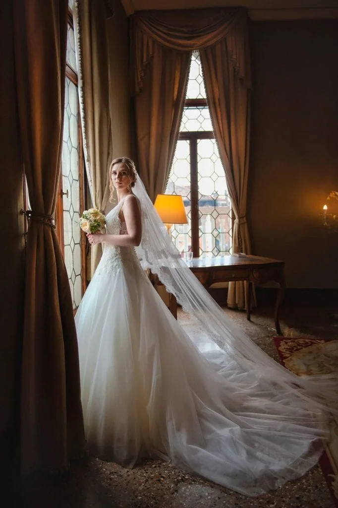 A bride standing elegantly in front of a large window at Ca’ Sagredo Palace in Venice wedding photography session.