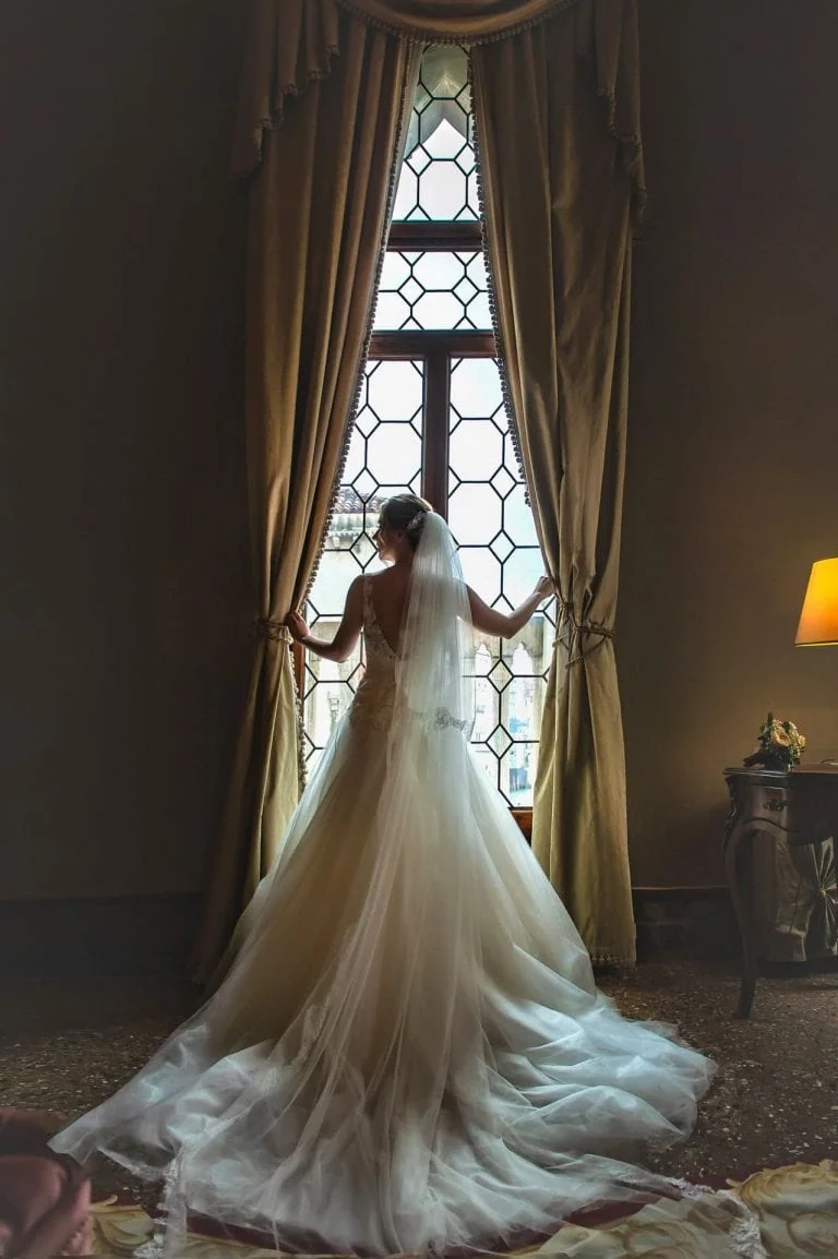 A bride standing elegantly in front of a large window at Ca’ Sagredo Palace in Venice – wedding photography Venice.