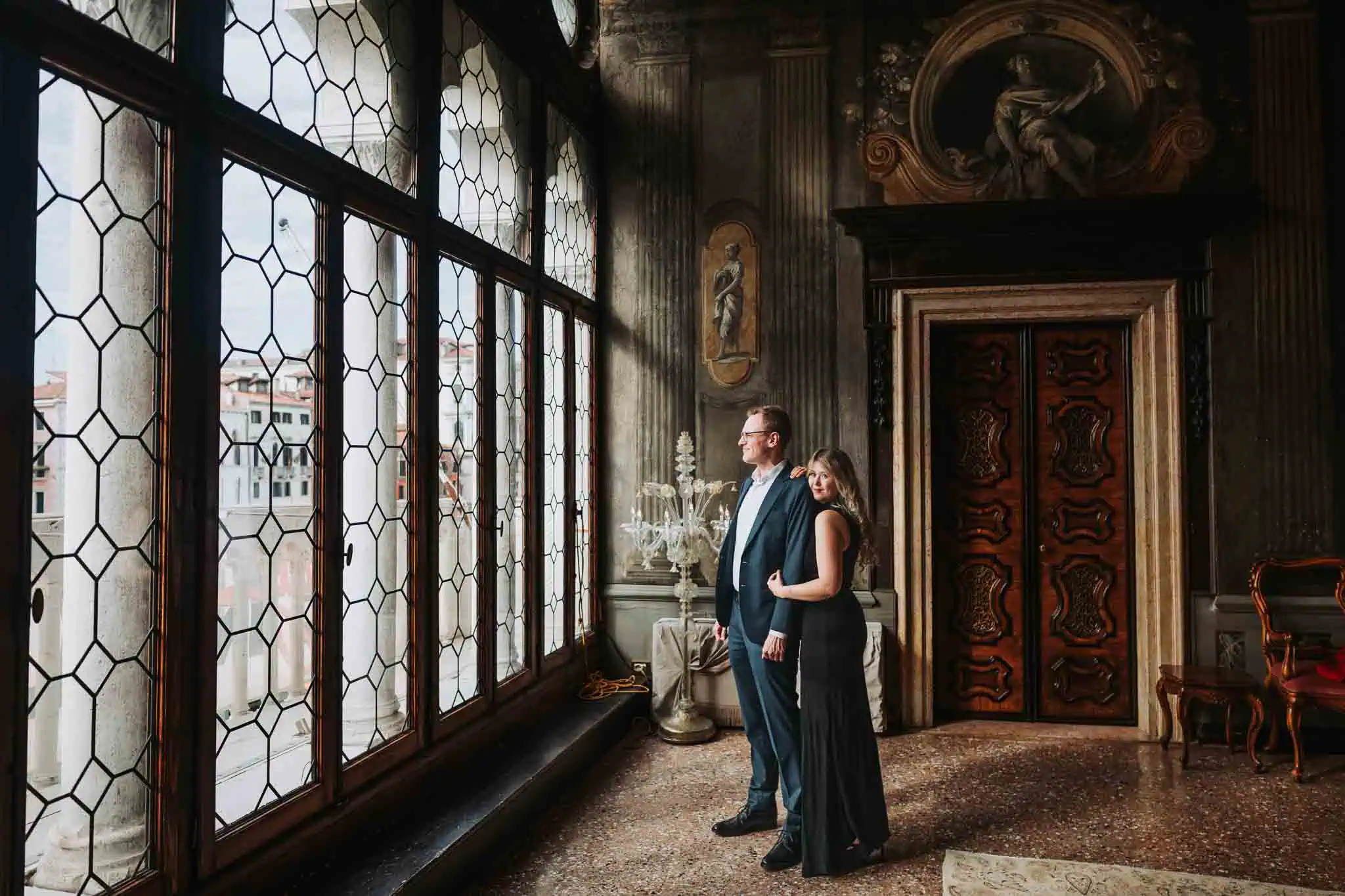 Elegant couple in formal attire inside historic Venetian building with ornate decor and large leaded glass windows.