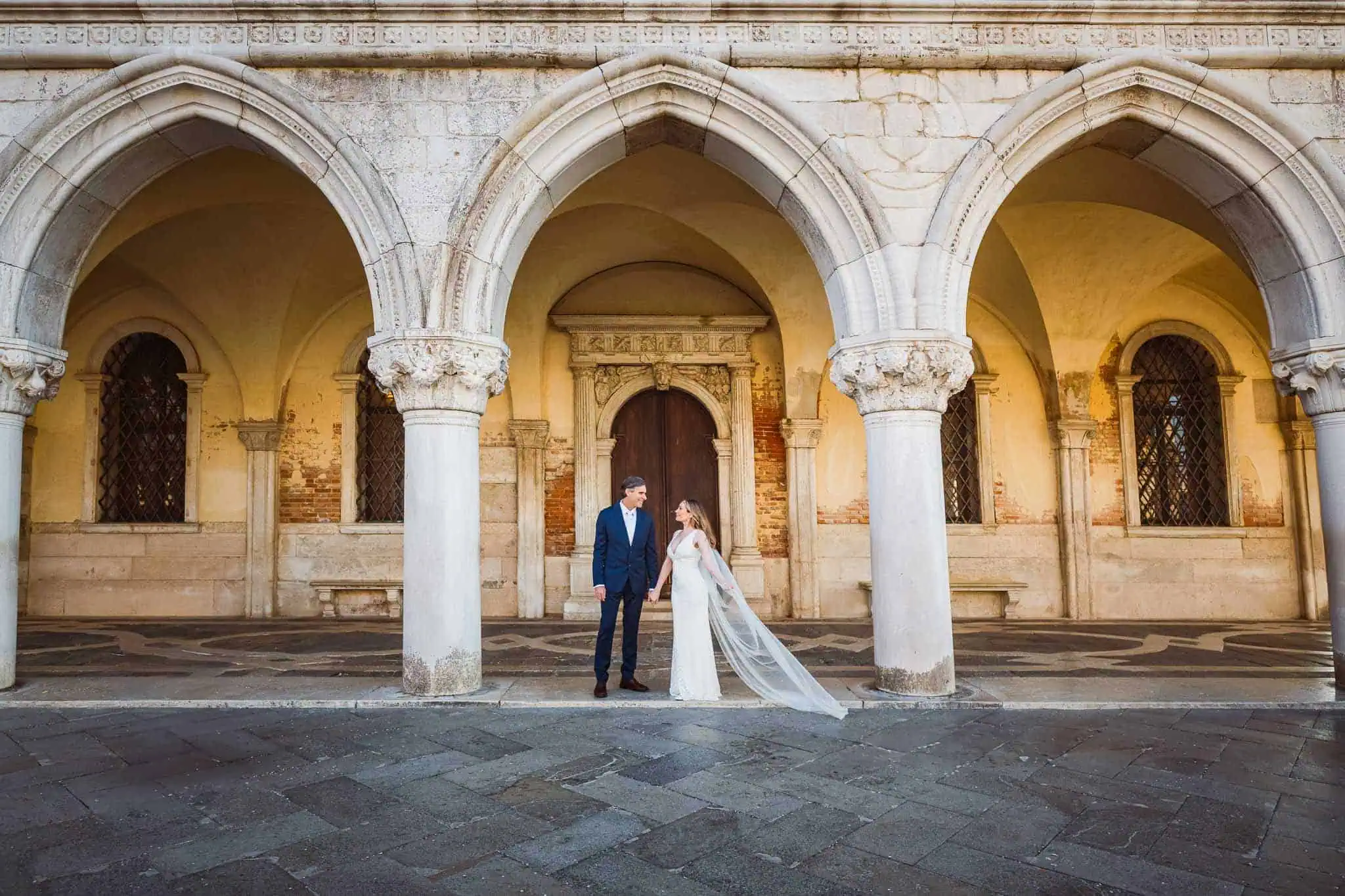 Elegant couple holding hands in Venice under historic arches, capturing romantic wedding moments.
