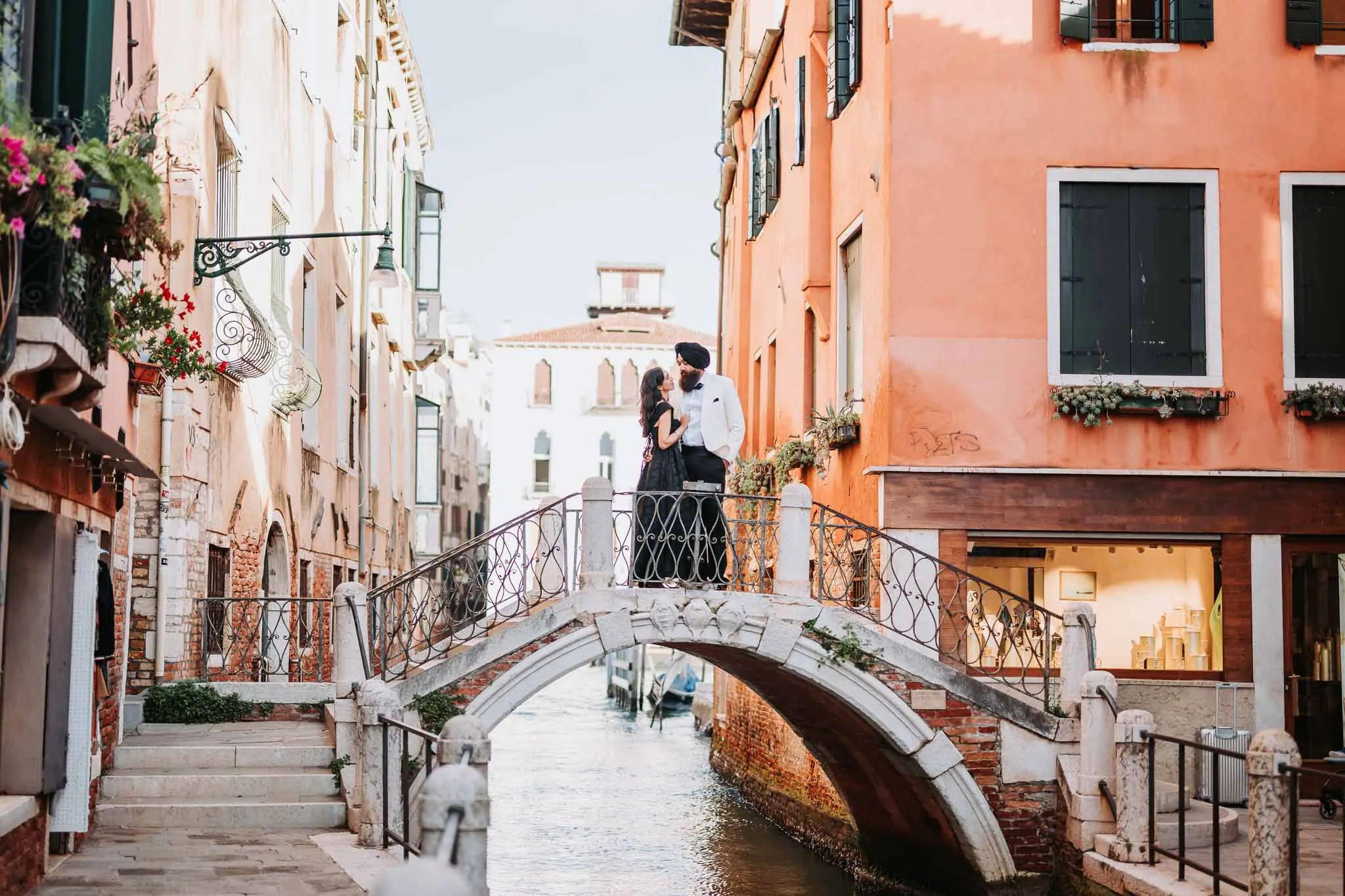 Romantic couple on a small Venetian bridge over a canal, surrounded by colorful historic buildings.