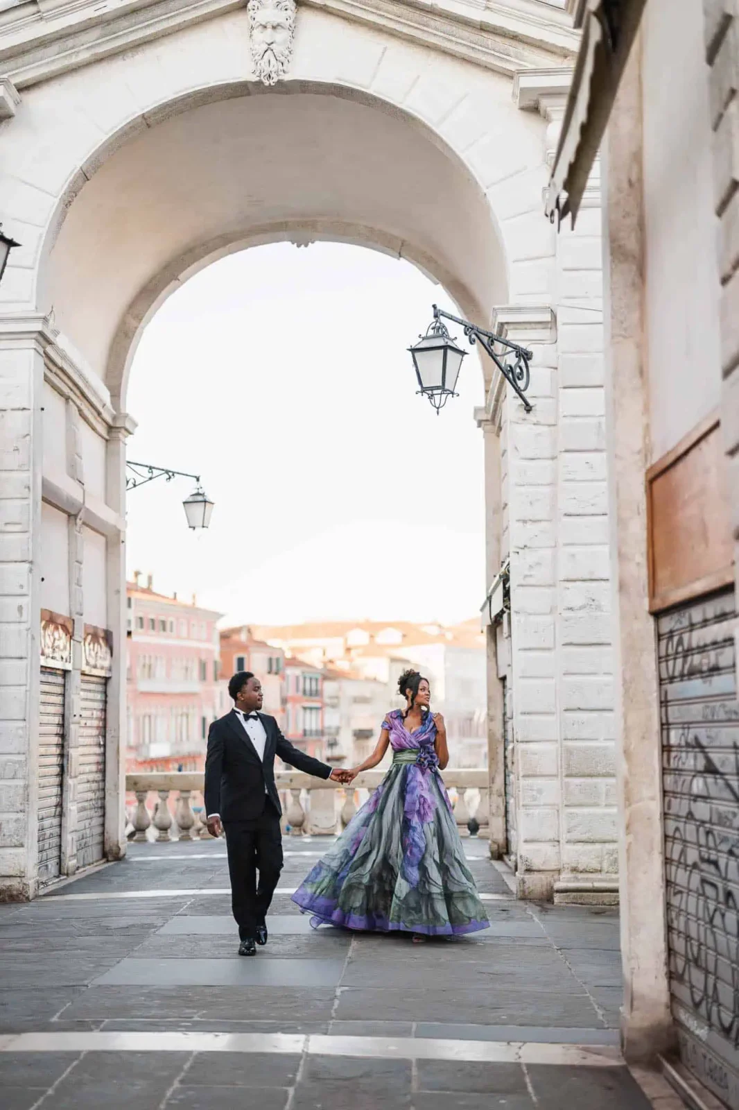 Elegant couple holding hands walking under Venice archway, romantic wedding proposal, engagement photo in Italy, artistic urban wedding shot.