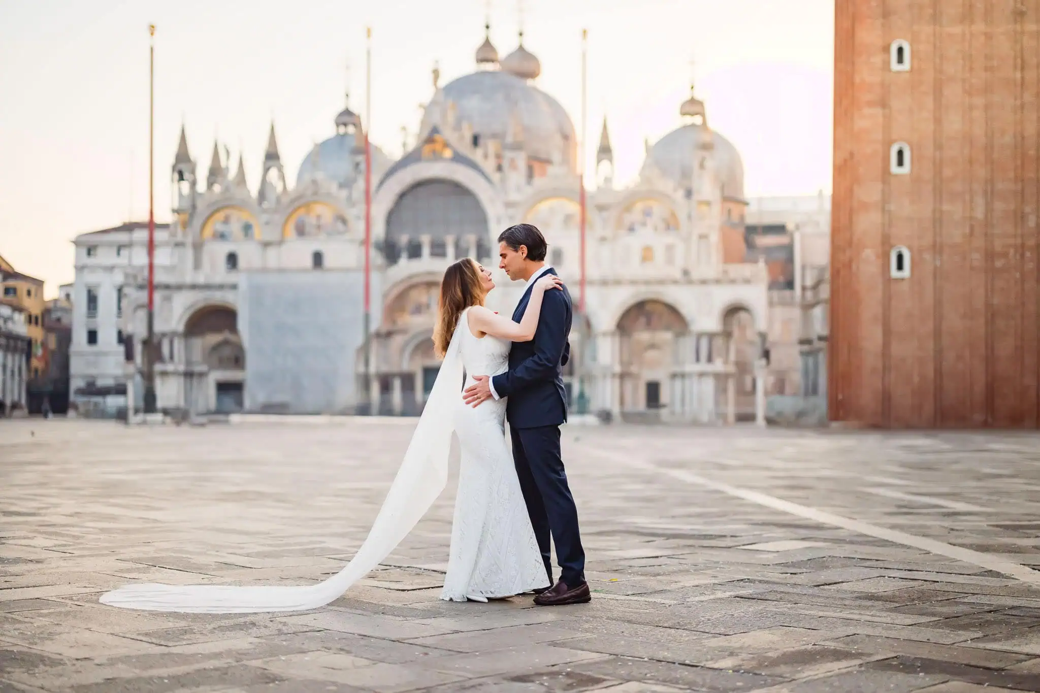Elegant couple in wedding attire embracing in Venice, with historic architecture in the background.