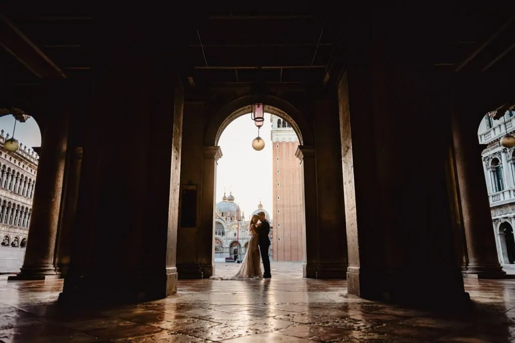 Bride and groom walk hand in hand through an empty Piazza San Marco, with the Bridge of Sighs in the background.