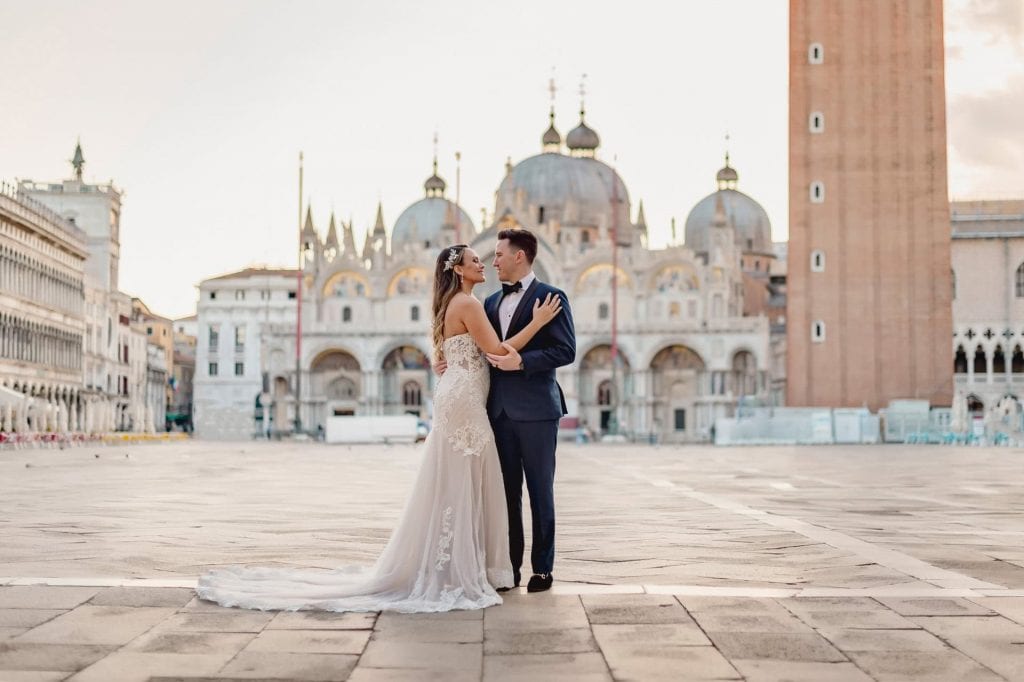 Bride and groom walk hand in hand through an empty Piazza San Marco, with the Bridge of Sighs in the background.