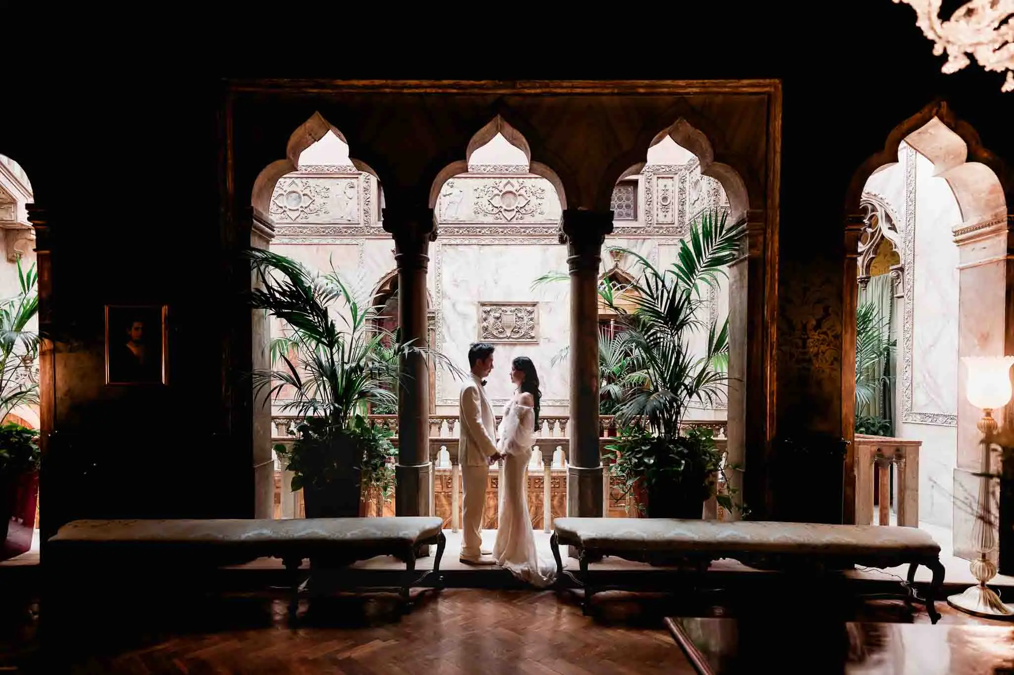Venice wedding couple holding hands in historic palace with ornate arches and lush plants.