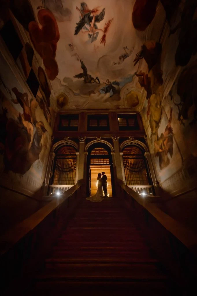 Newlyweds pose on a grand staircase with angel sculptures and frescoed walls in a Venetian palace.