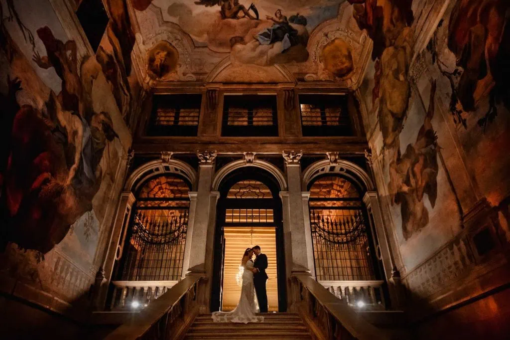 Newlyweds pose on a grand staircase with angel sculptures and frescoed walls in a Venetian palace.
