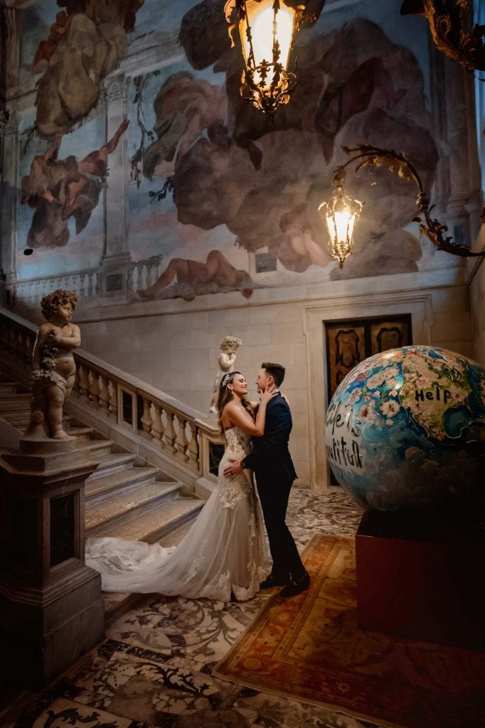 Newlyweds pose on a grand staircase with angel sculptures and frescoed walls in a Venetian palace.
