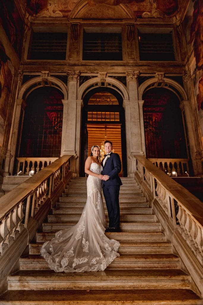 Newlyweds pose on a grand staircase with angel sculptures and frescoed walls in a Venetian palace.