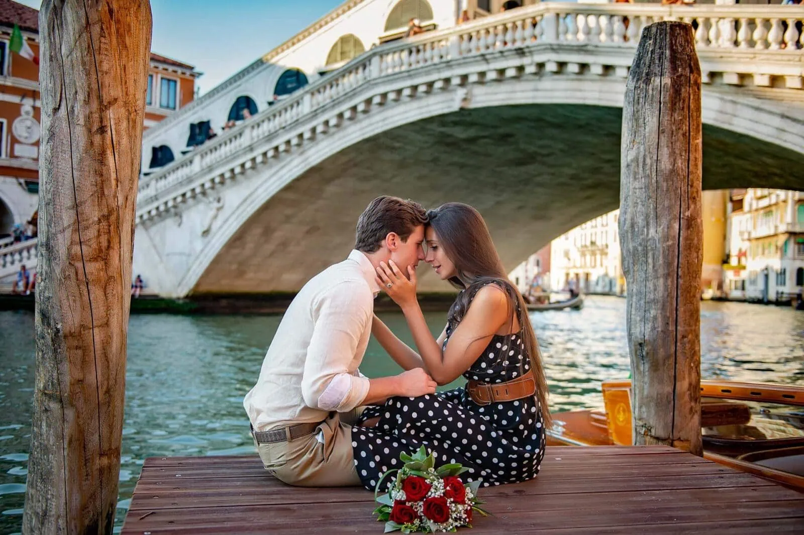 Engagement photos in Venice featuring a couple embracing with a bouquet of roses in front of Rialto Bridge.