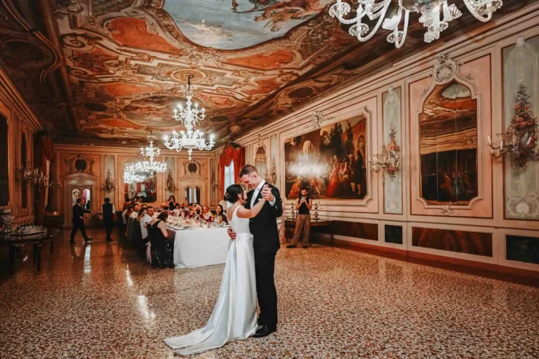Elegant couple sharing a first dance in a luxurious, ornately decorated ballroom with chandeliers and classical artwork.