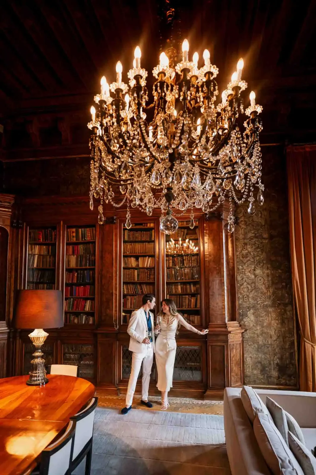 Elegant couple in sophisticated ivory attire inside a luxurious, vintage-style room with grand chandelier and wooden bookshelves.