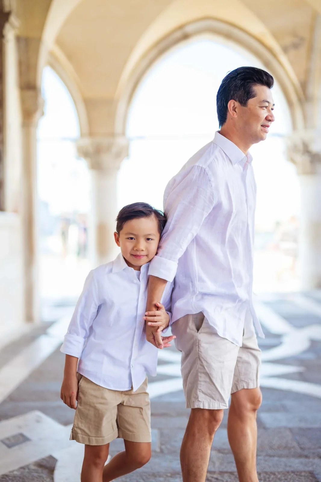 A parent and child holding hands at Venice, outdoors during daytime, wearing white shirts and beige shorts.