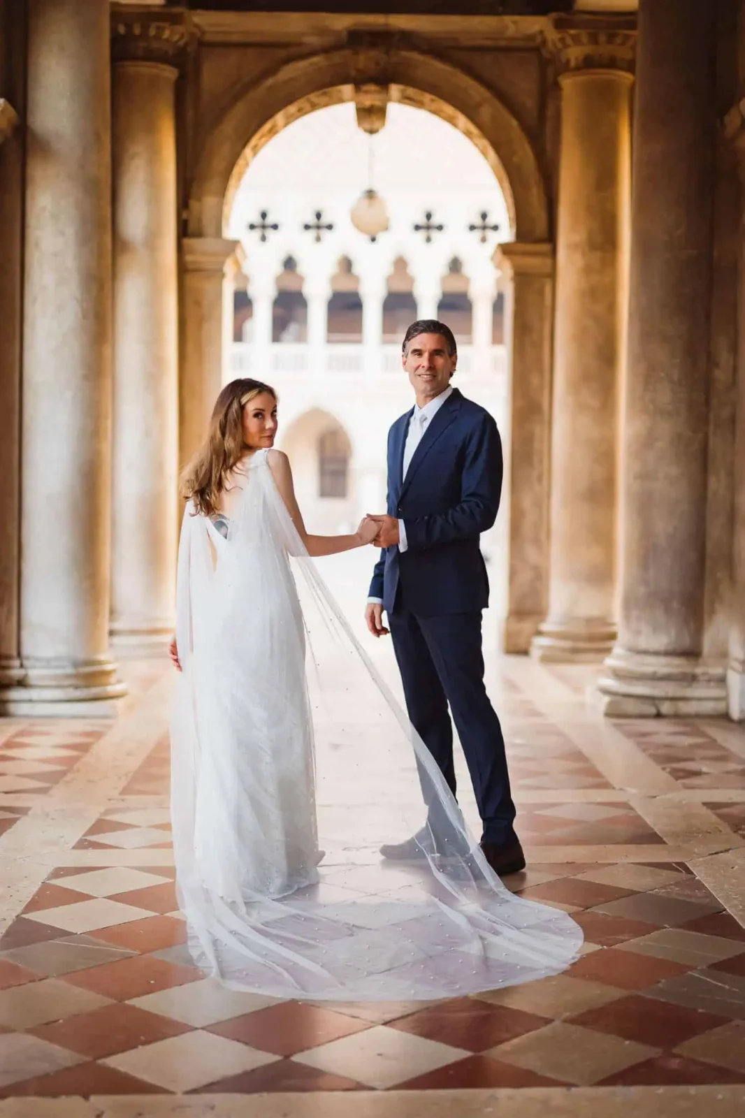 Elegant couple in wedding attire holding hands at Venice historic palace.