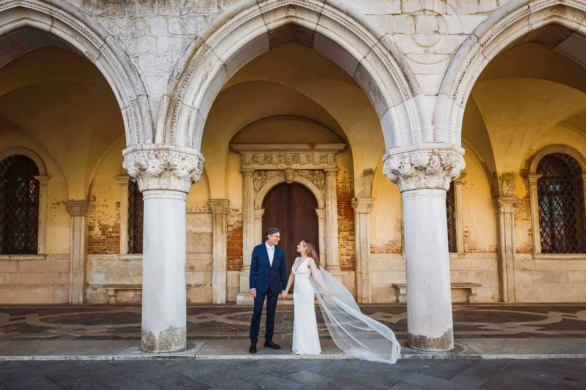 Elegant wedding couple holding hands under historic Venetian arches, romantic Venice wedding photography.