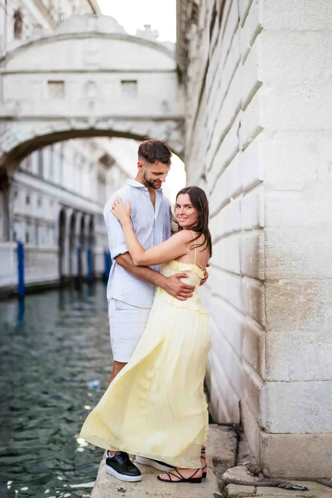 Romantic couple embracing in Venice canal with historic bridge backdrop, candid love moment.