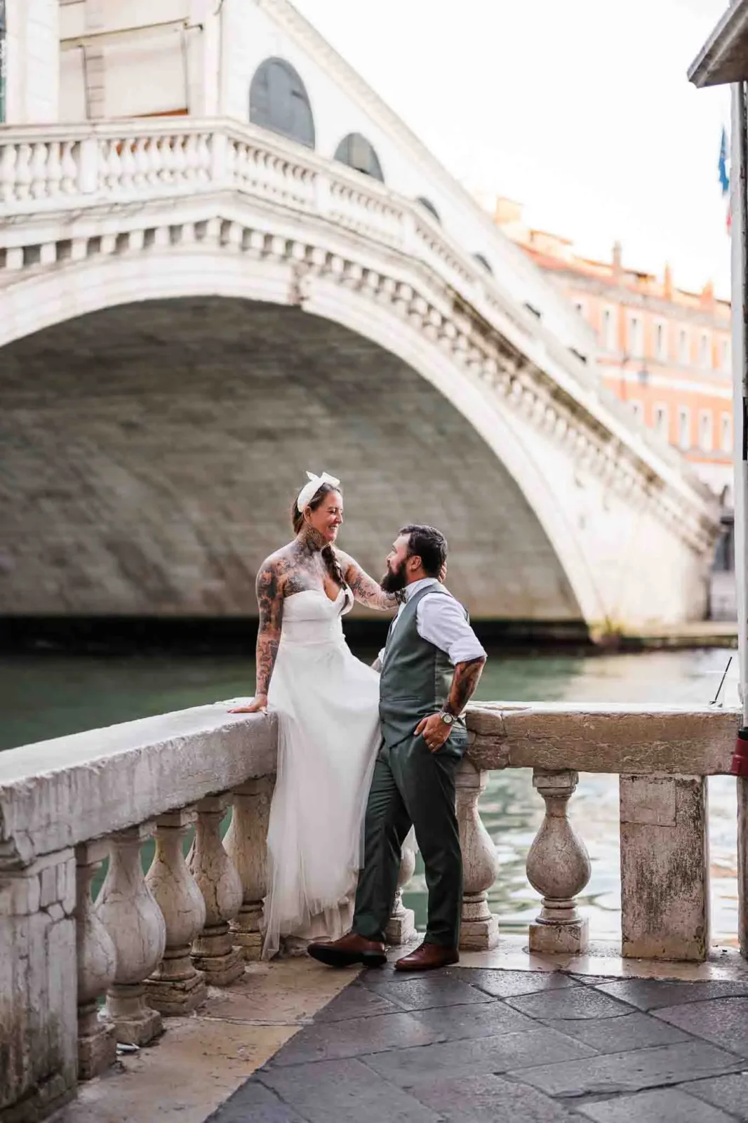 1. Tattooed bride in wedding dress laughing with groom by Venice canal under historic bridge.
