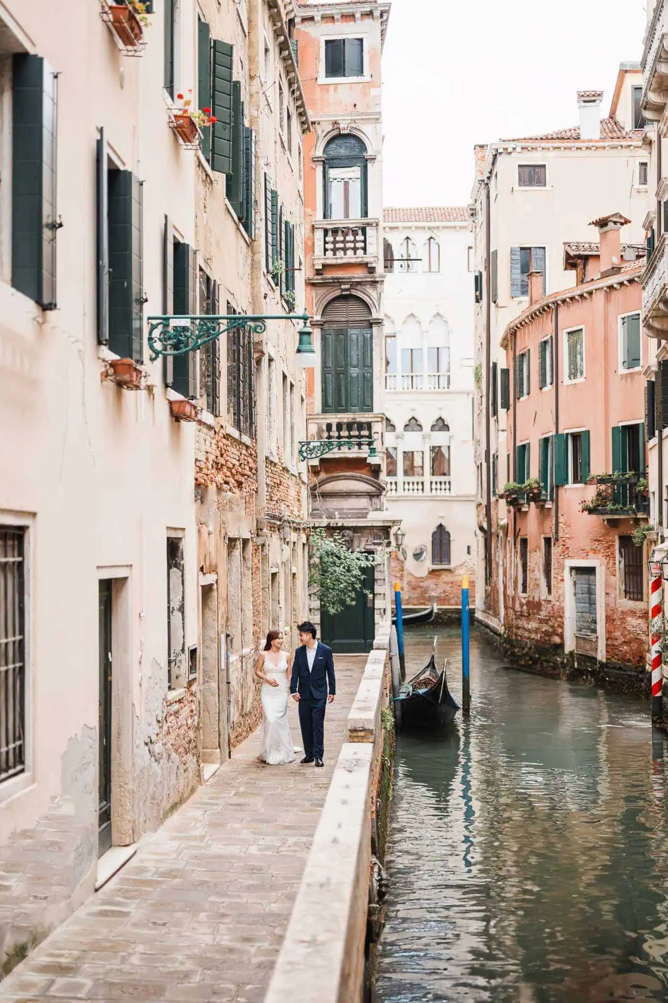 Charming couple walking along Venice canal in wedding attire, romantic backdrop.