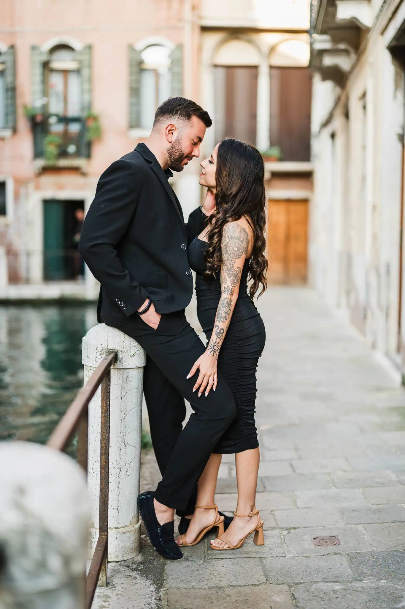 Elegant couple in black attire sharing a romantic moment on a Venice canal bridge.