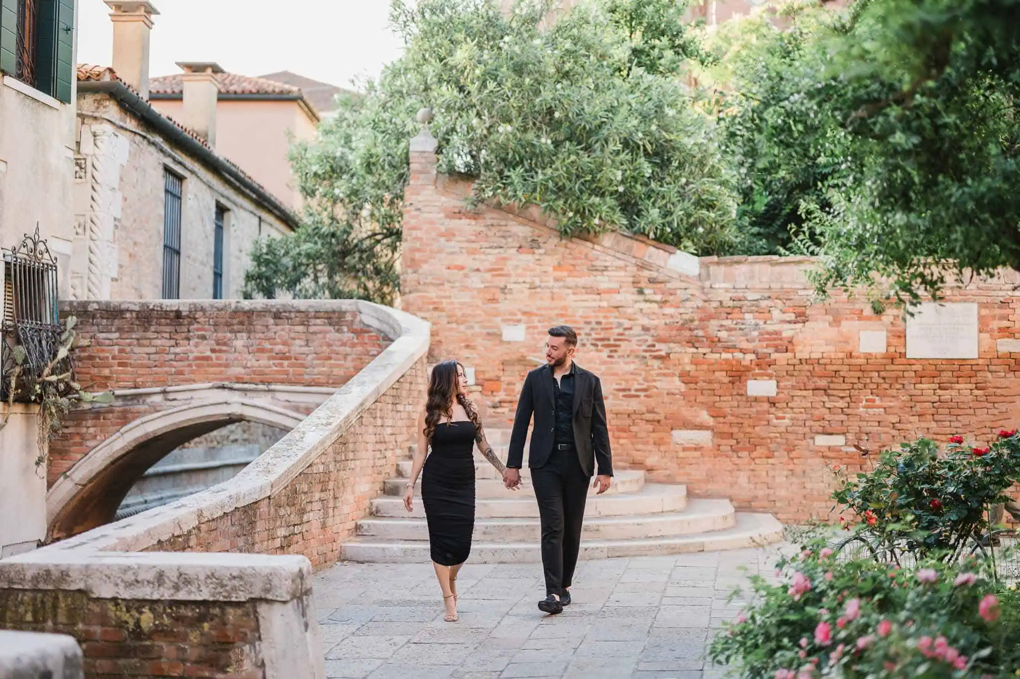 Romantic couple holding hands in Venice, beautiful historic bridge and lush greenery in the background.