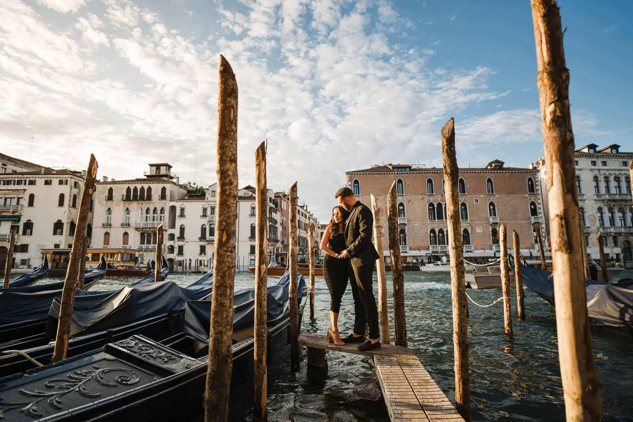 Romantic couple embracing on Venice gondola dock during sunset, picturesque cityscape backdrop.