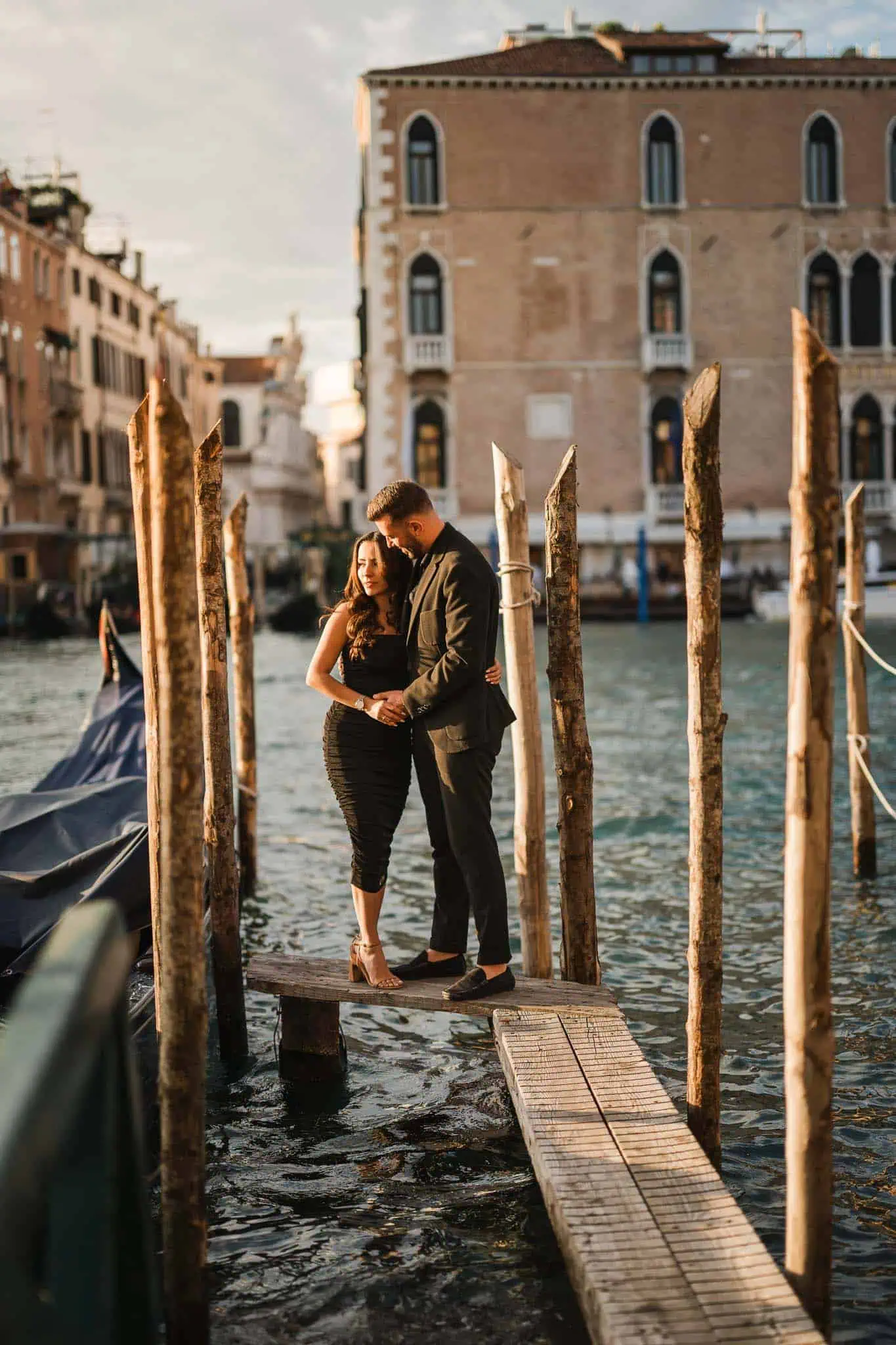 Romantic couple in elegant attire sharing a moment on Venice gondola dock at sunset.