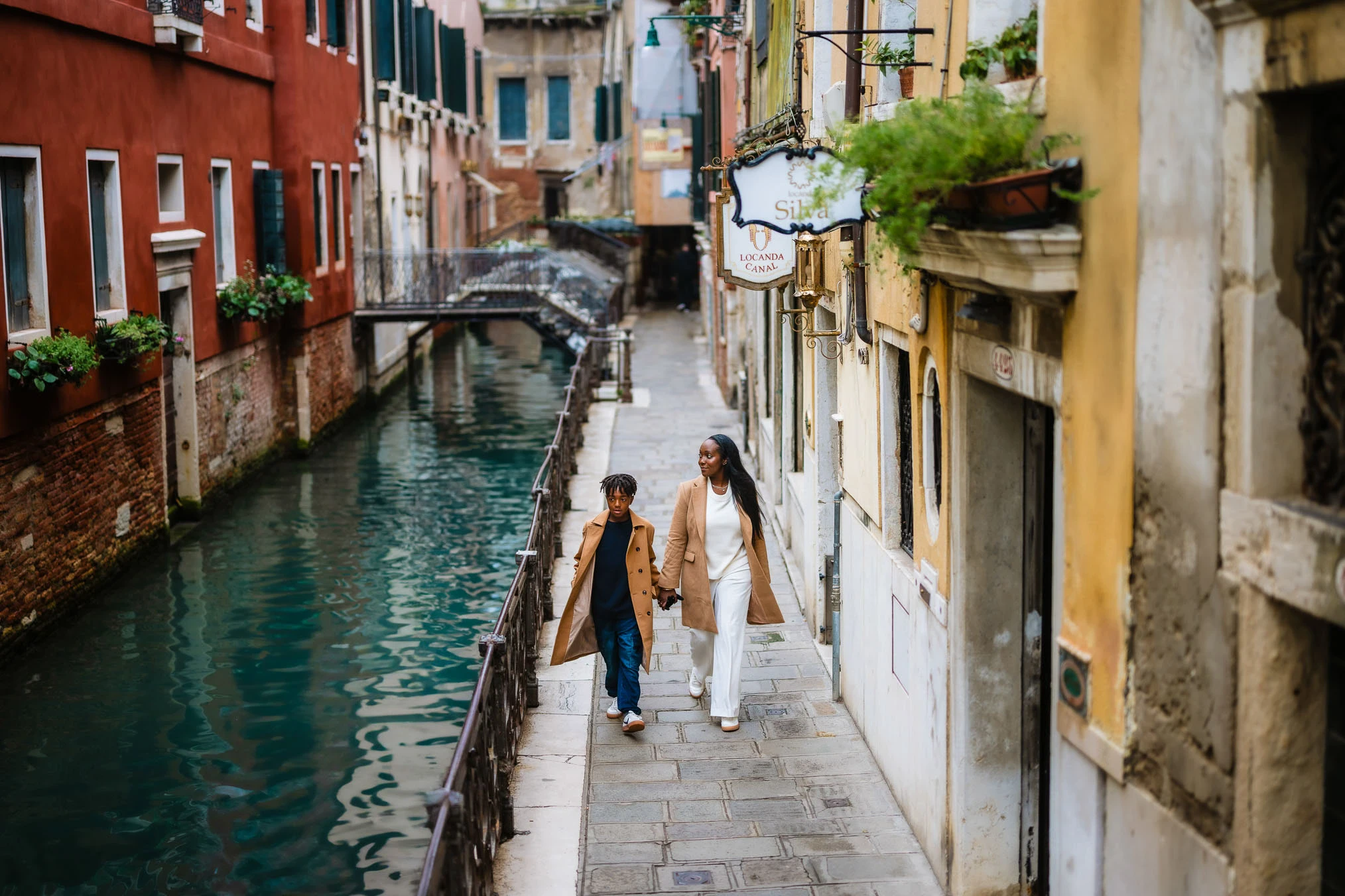 Elegant woman and child strolling along Venice canal in fashionable coats, charming scenery, romantic atmosphere.