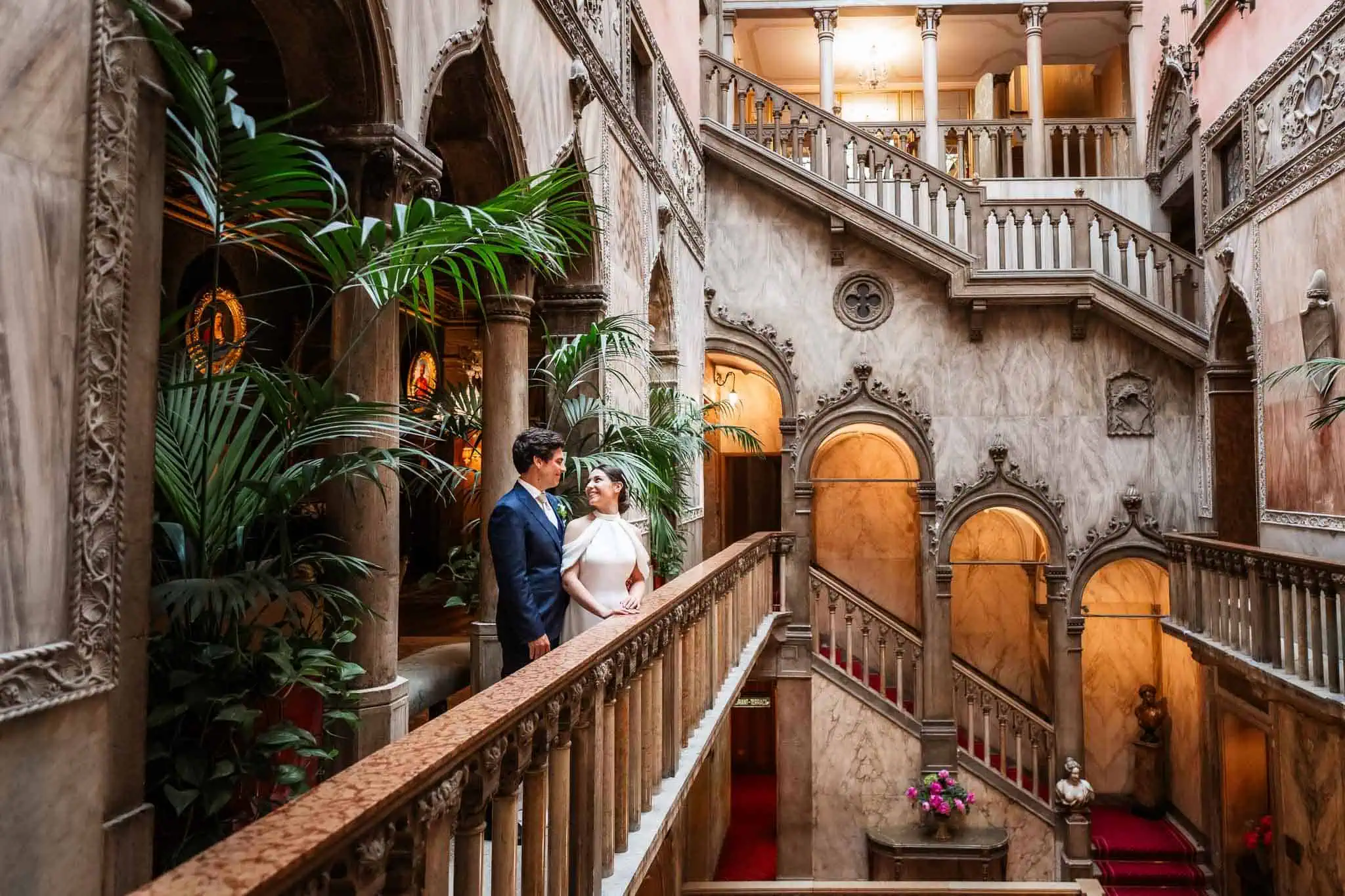 Elegant couple in wedding attire inside a grand Venetian palace staircase.