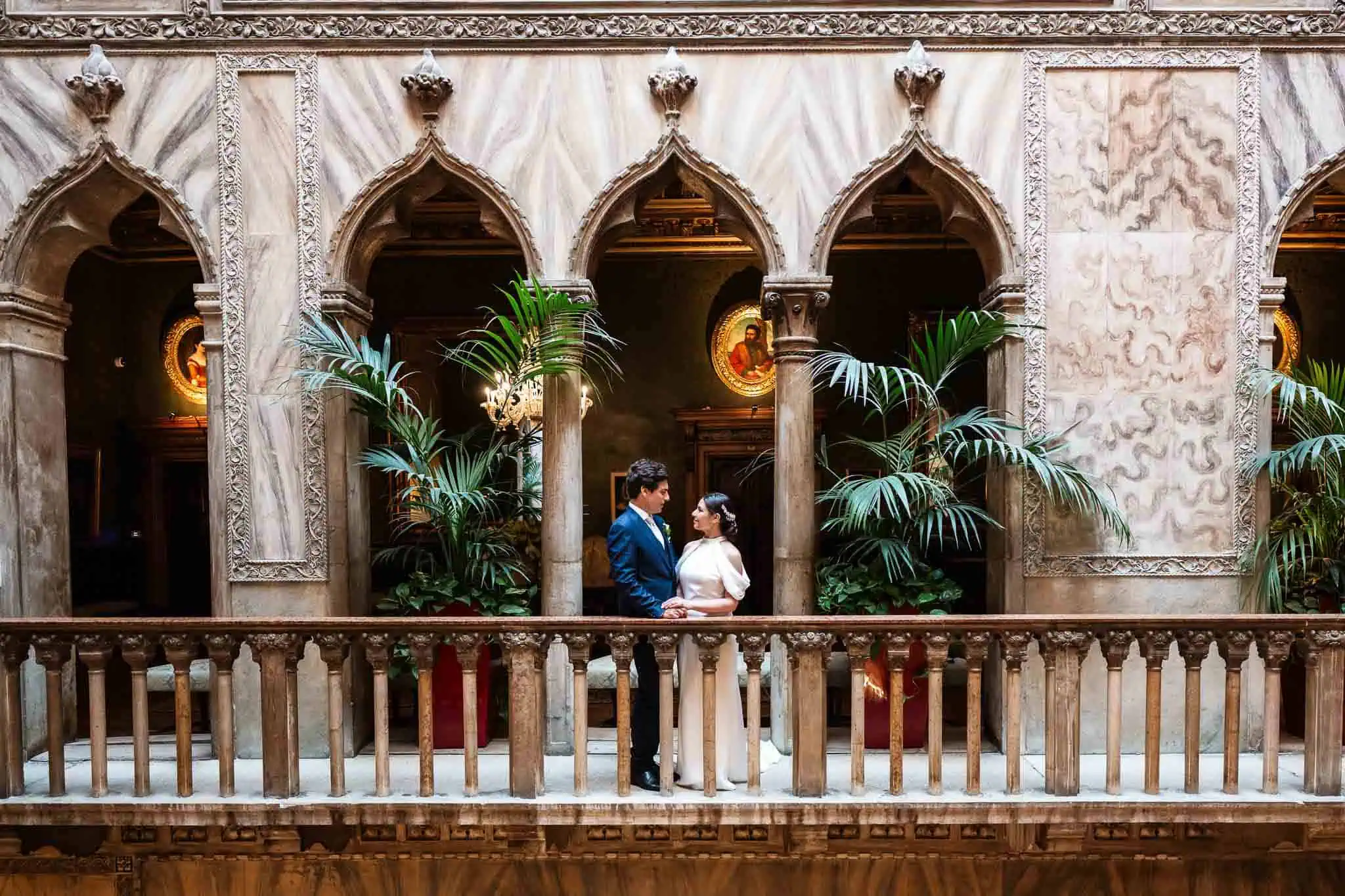 Elegant couple holding hands on a Venetian balcony with ornate arches and lush greenery, capturing romantic wedding moments.
