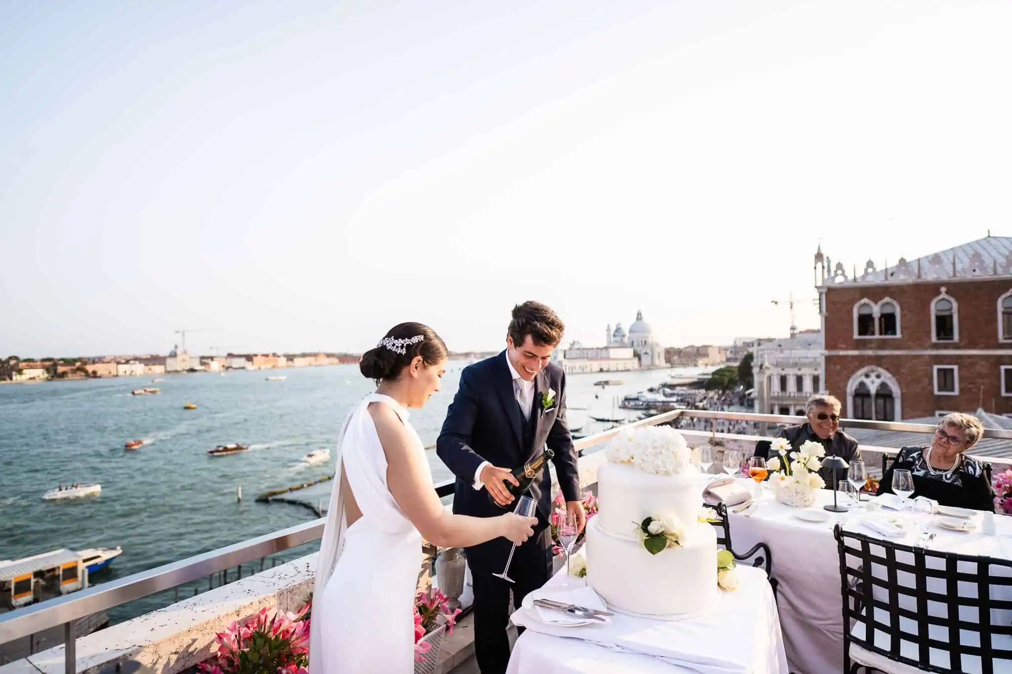 Elegant couple cutting wedding cake on Venice rooftop with canal view, historical architecture, and scenic sunset.