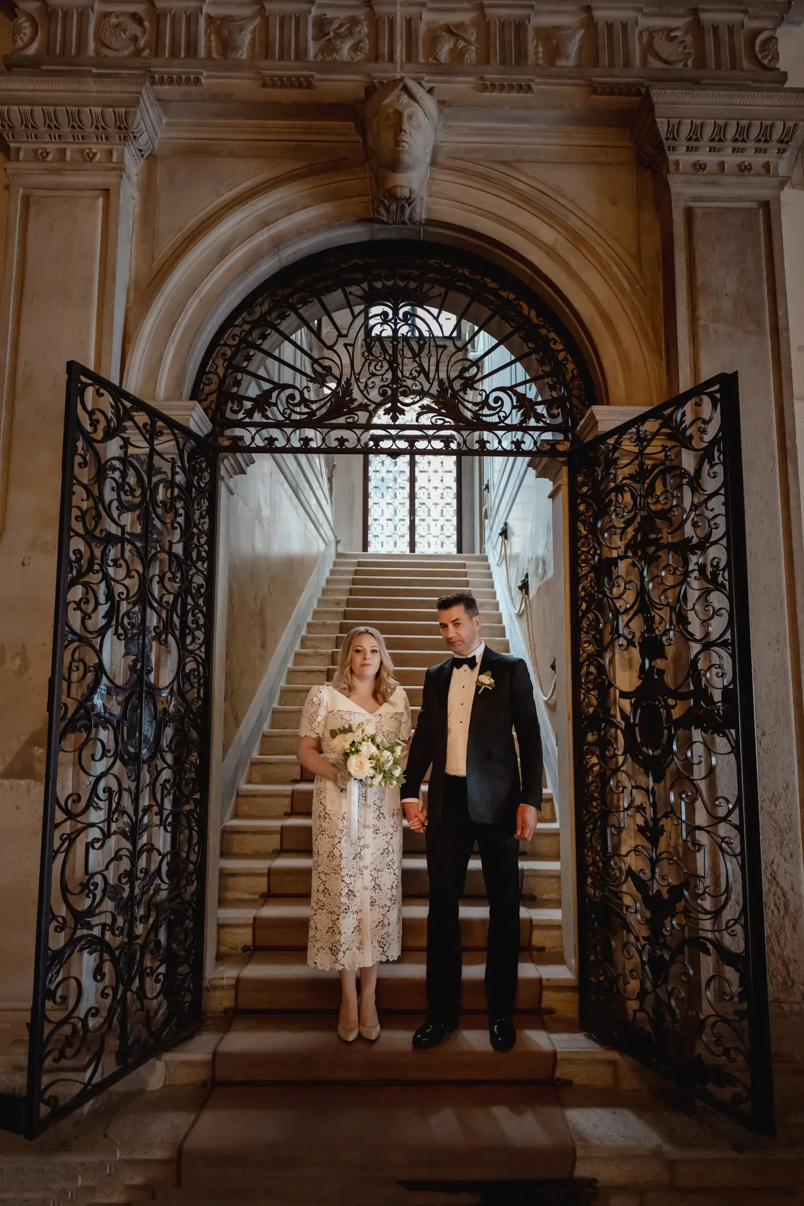 Elegant couple stands on grand staircase inside historic building, celebrating their elopement.