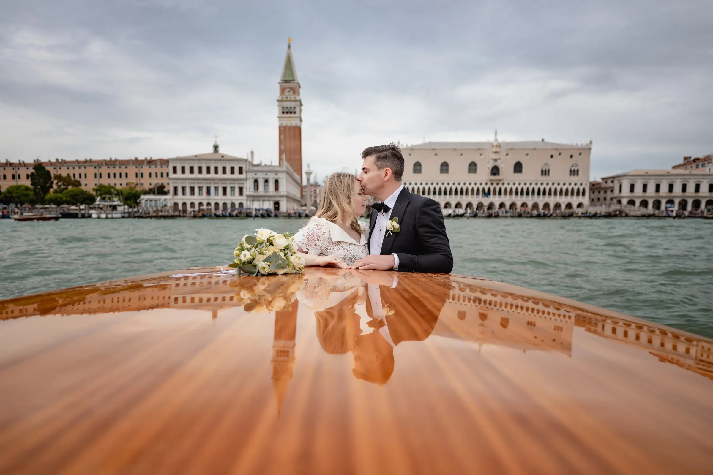 Elegant couple sharing an intimate moment on a wooden boat in Venice, with historic architecture in the background.