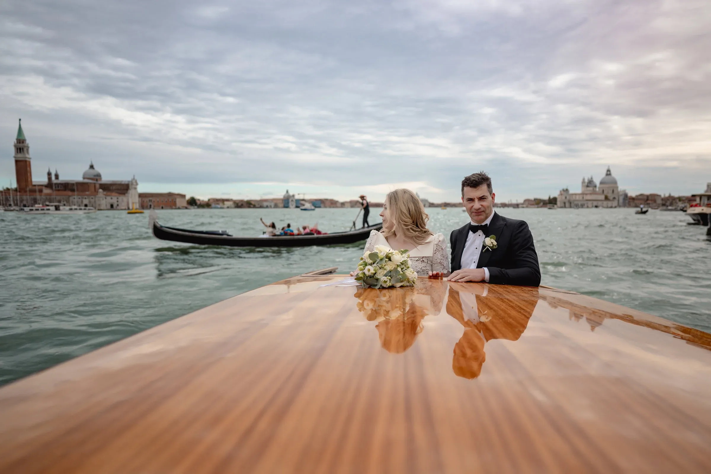 Romantic couple on a wooden boat during an elopement in Venice, with iconic cityscape and gondolas in the background.