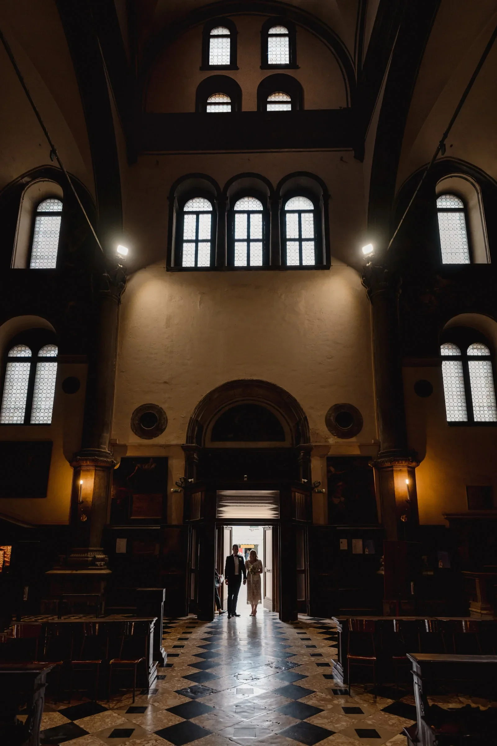 Elegant couple standing at church entrance with high arched windows and dramatic lighting.
