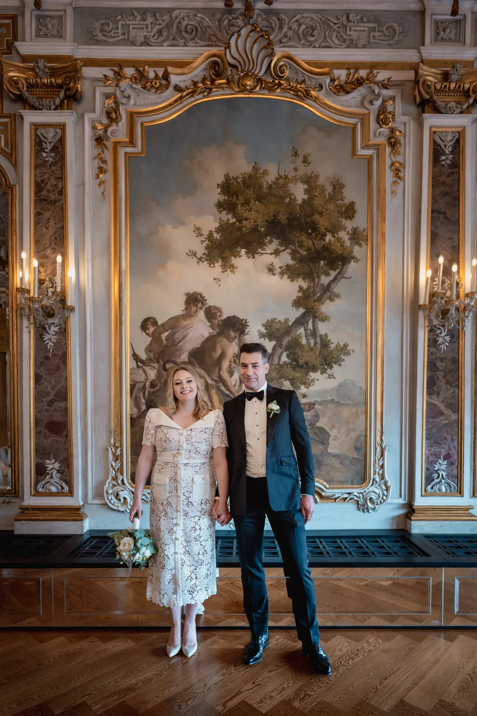 Elegant couple holding hands during their elopement inside a grand, ornate hall with classical artwork and luxurious decor.