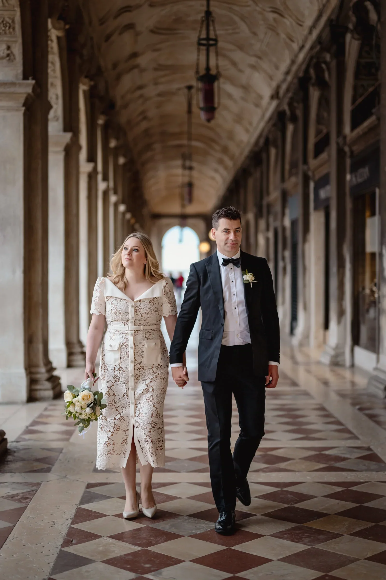 Elegant couple in vintage attire walking through historic archway corridor.