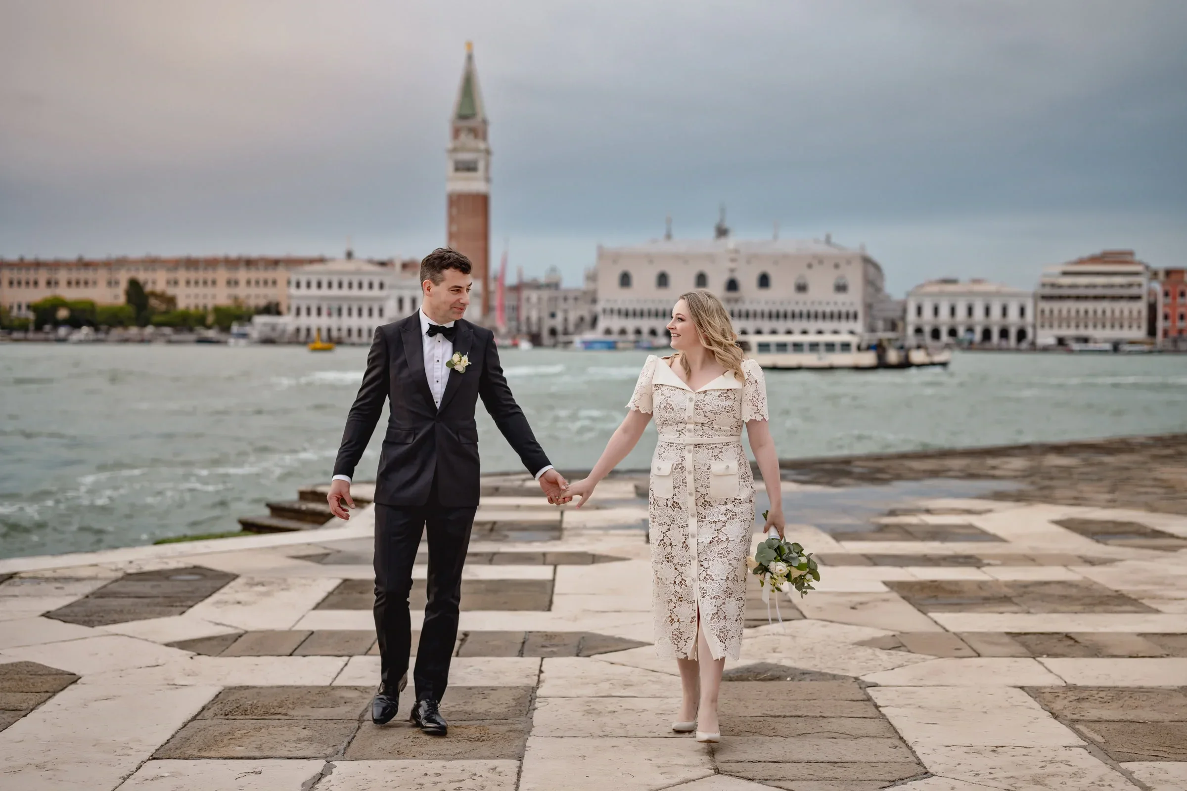 Elegant couple in wedding attire walking hand in hand by the water in Venice, with historic architecture in the background.