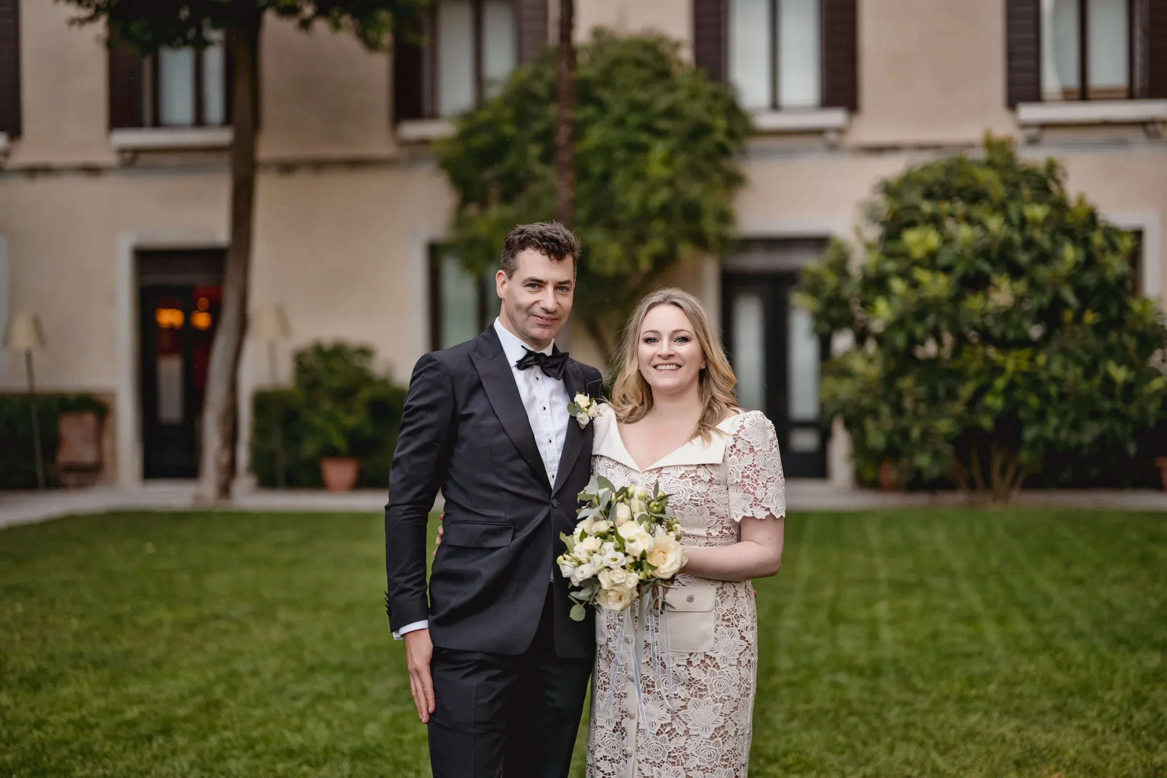Elegant couple in wedding attire celebrating outdoors amid greenery and a charming building backdrop.