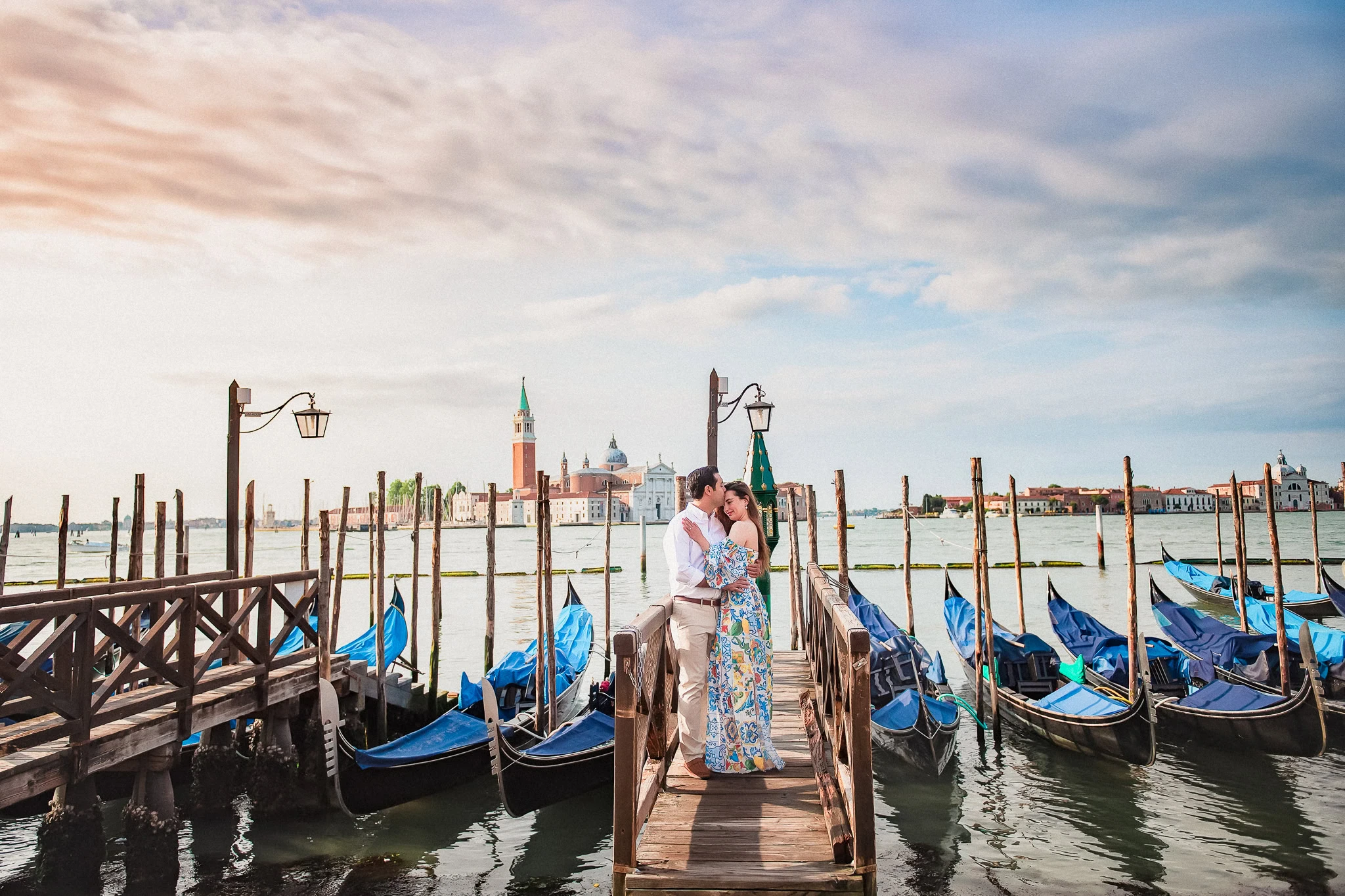 Romantic Venice engagement photo on gondola dock with couples kissing, scenic city skyline, and colorful sky.