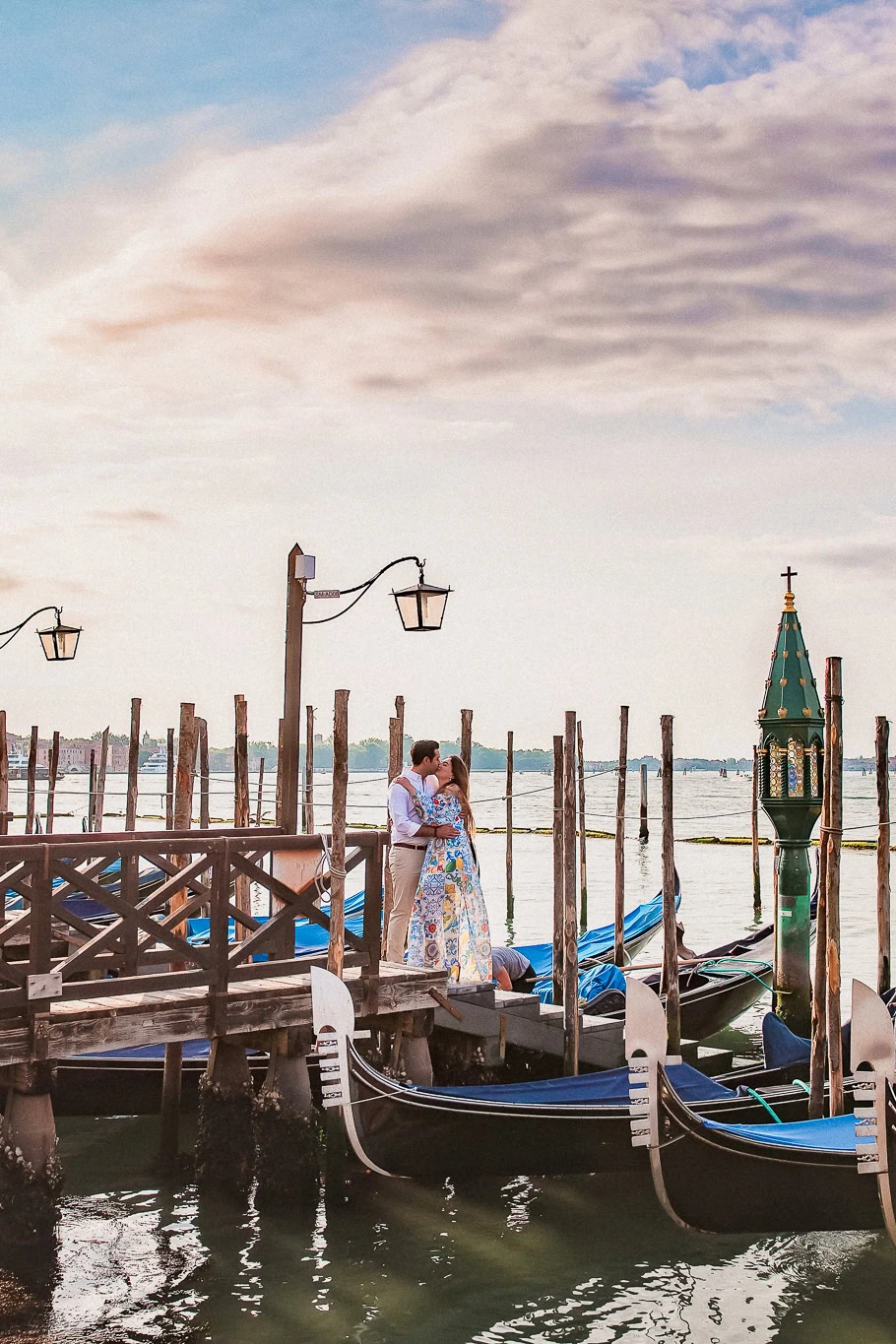 Romantic Venice couple kissing at sunset by gondolas on water, scenic sky background.