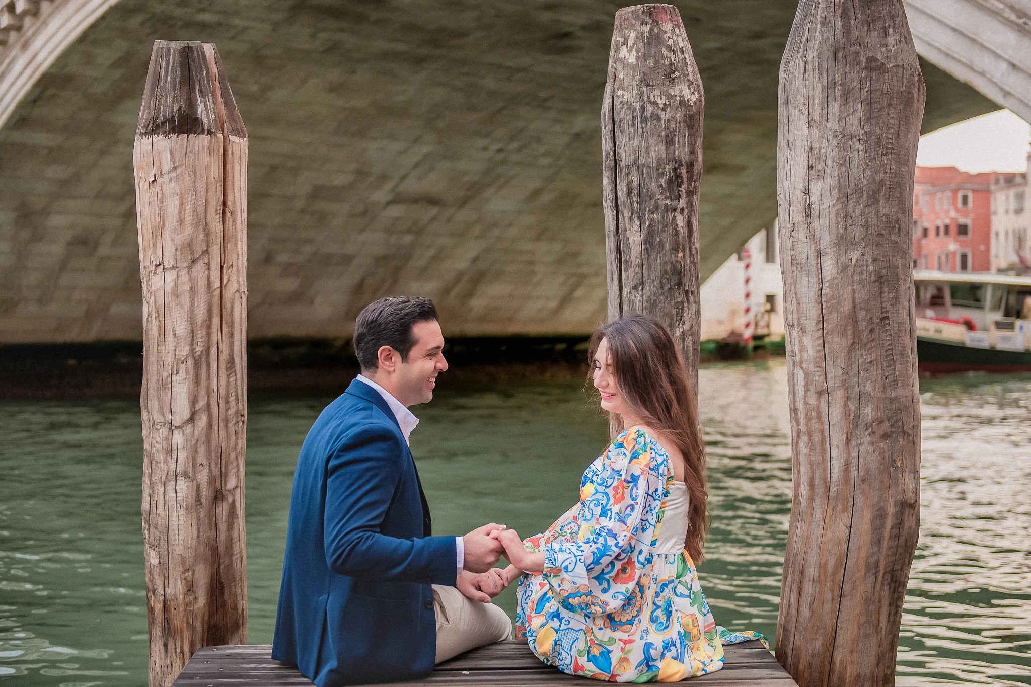 Romantic Venice proposal photo of couple sitting on dock by canal under bridge, holding hands.