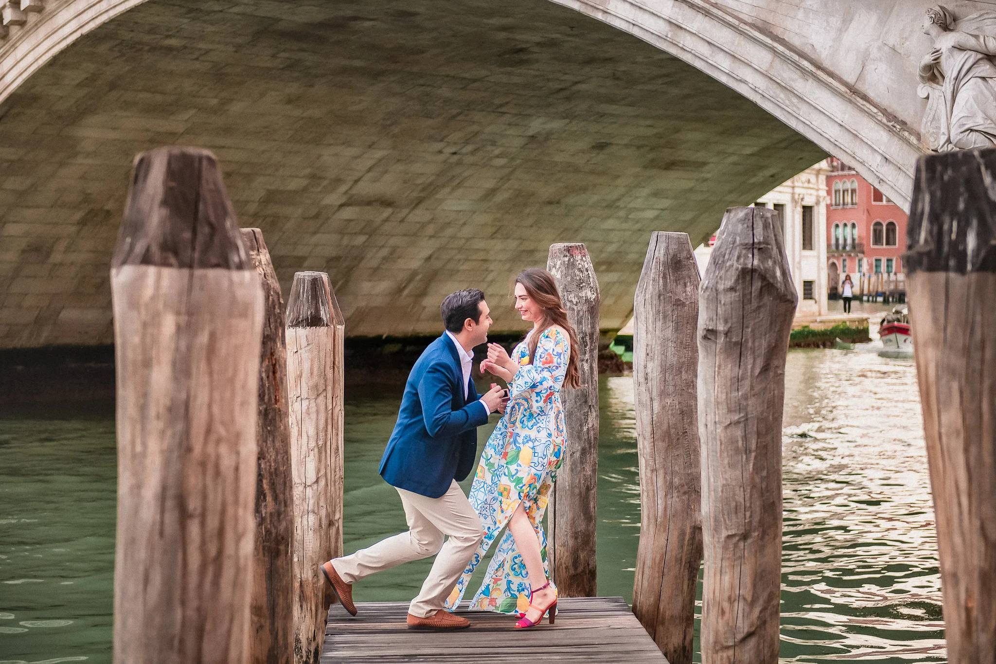 Romantic proposal on Venice gondola dock under historic bridge with colorful buildings in background.